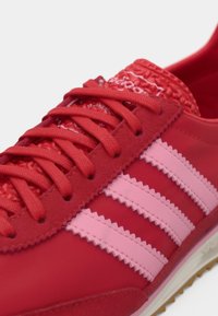 Baskets rouges avec une tige texturée, des accents roses et trois bandes roses. Les lacets sont également rouges, avec une semelle en caoutchouc pour l'adhérence.