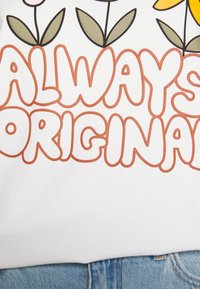 Vit bomull t-shirt med djärv, rundad orange text som säger "ALWAYS ORIGINAL" med blommönster i dämpade färger ovanför.