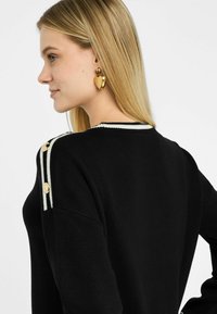 Zwarte trui met witte gestreepte afwerking bij de halslijn en schouder, met decoratieve gouden knopen op de schouder. Soepele textuur.