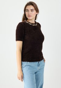Brauner Pullover mit kurzen Ärmeln und rundem Ausschnitt, verziert mit dekorativen Perlen am Kragen. Weiche Textur, leicht tailliertes Design.