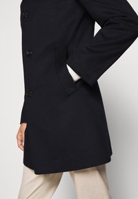 Cappotto blu navy con superficie liscia, chiusura con bottoni, tasche laterali e leggero taglio svasato, abbinato a pantaloni beige.