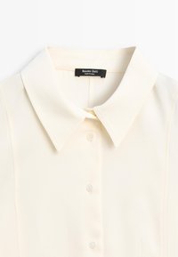 Cremefarvet skjorte med klassisk krave, der har fem knapper langs forsiden og en blød, glat stofstruktur. Etiket synlig ved kraven.