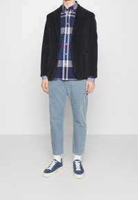 Zwarte corduroy blazer, marineblauw en geruit overhemd met knopen, lichtblauwe gestreepte broek en blauwe sneakers met witte zolen en veters.