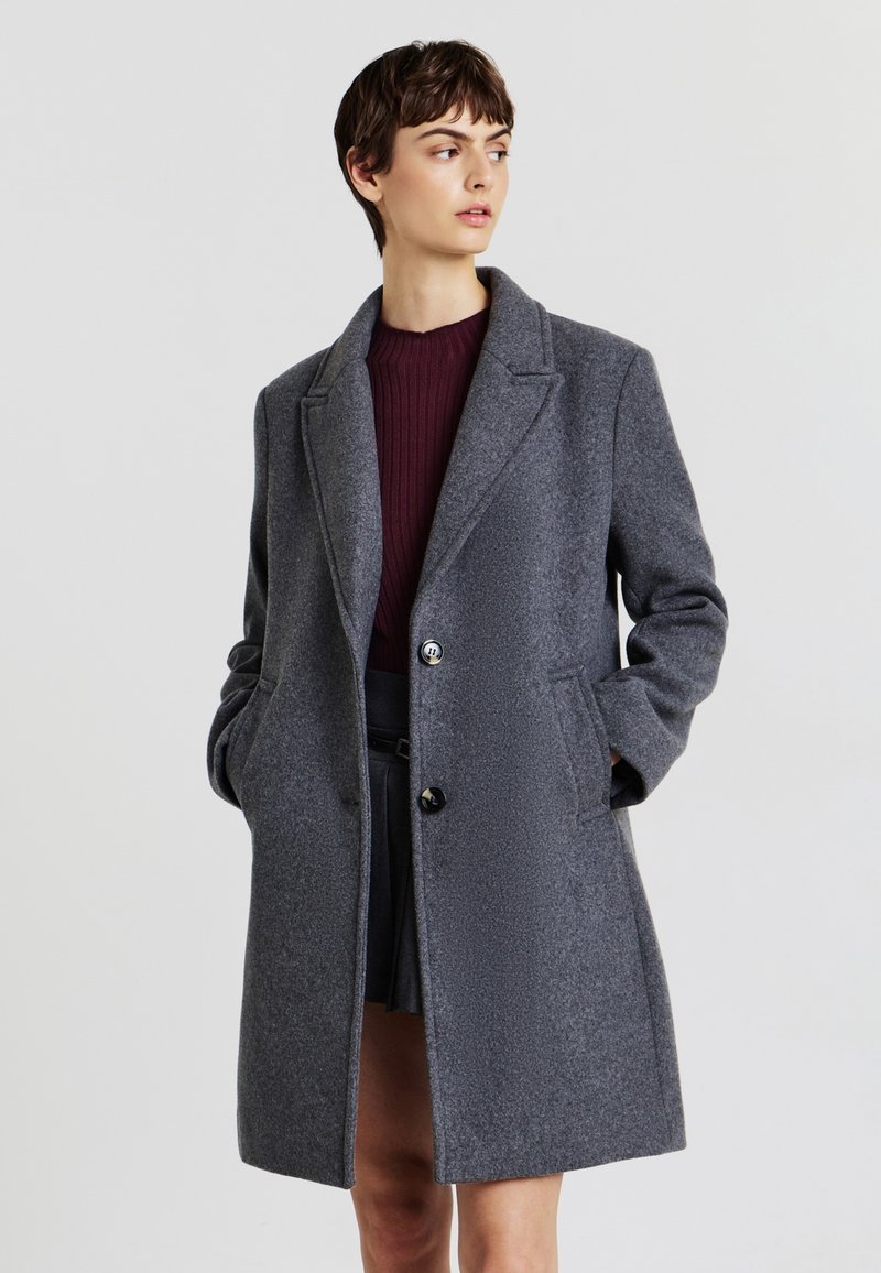 Cappotto grigio in lana con colletto a punta, chiusura con un bottone singolo e tasche laterali. Il tessuto ha una texture sottile e una finitura liscia.