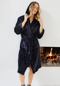 Robe de bain en peluche noire avec capuche, dotée d'une ceinture, de manches longues et de poches latérales, se tenant devant une cheminée.