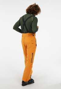 Oranje skibroek met ritszakken, verstelbare banden en een strak design. Draagt met een groene lange mouw top en zwarte skischoenen.