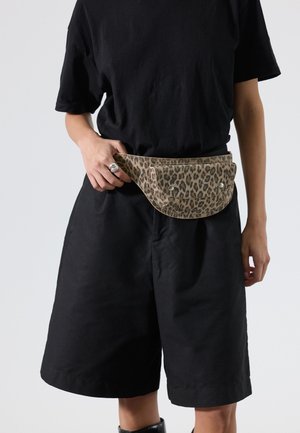 Persoon die zwarte oversized shorts en een t-shirt draagt met een luipaardprint heuptasje vastgemaakt rond de taille, die het tasriem vasthoudt.