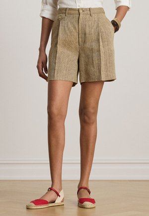 Žena v béžových vysokých šortkách s rybej kosti, bielej dlhoruká blúzka s vyhrnutými rukávmi, červených espadrilách s remienkom okolo členka a hnedom náramku.