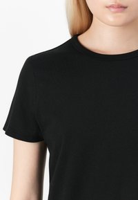 Personne blonde portant un t-shirt noir uni à manches courtes et col rond, sur fond blanc, vue de la moitié de la poitrine vers le haut.