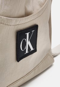 Mochila de tela beige con superficie texturada, un parche rectangular negro con el logo blanco "cK" y un bolsillo lateral.