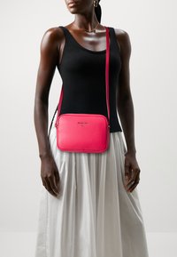 Borsa a tracolla rettangolare di colore rosa acceso, in pelle testurizzata, con logo argentato e tracolla regolabile. Indossata con un top in cotone nero e una gonna bianca.