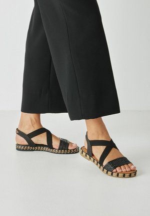 Zwarte geweven sandalen met gekruiste bandjes, gestructureerd bovenwerk en een geweven zool van jute en rubber. Vlak ontwerp voor comfort en stabiliteit.