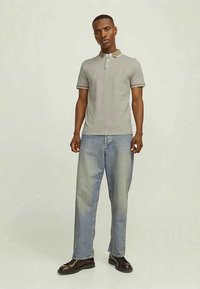 Homem em pé, vestido com uma camisa polo cinza claro, jeans azuis largos e sapatos castanhos escuros, contra um fundo claro e simples.