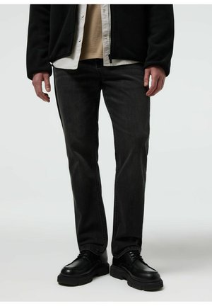 Mężczyzna ubrany w czarne proste jeansy, czarne masywne buty sznurowane, beżową koszulę, biały podkoszulek i czarną kurtkę, stojący na jednolitym tle.