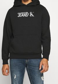 Felpa nera con il logo bianco "Calvin Klein Jeans cK" indossata con jeans blu strappati da una persona su uno sfondo semplice.