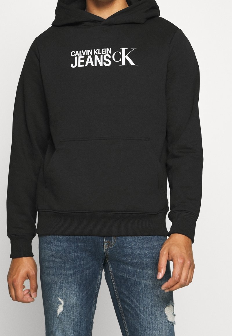 Felpa nera con il logo bianco "Calvin Klein Jeans cK" indossata con jeans blu strappati da una persona su uno sfondo semplice.