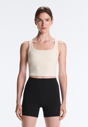 Mladá žena nosiaca béžový bezrukávový crop top a čierne šortky s vysokým pásom stojaca na jednofarebnom svetlom pozadí.
