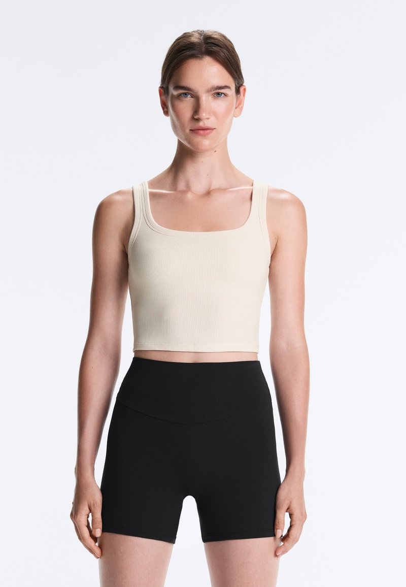 Jeune femme portant un crop top beige sans manches et un short noir taille haute, debout devant un fond clair uni.
