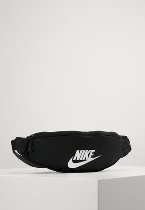 Schwarze Nike Gürteltasche mit weißem Logo, Reißverschluss und verstellbarem Riemen, auf einer weißen Fläche vor neutralem Hintergrund dargestellt.
