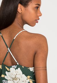 Vestido floral verde com um tecido texturizado, apresentando finas alças cruzadas nas costas e um decote nas costas. Exibe uma tatuagem no braço superior.
