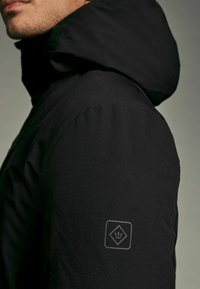 Juodas gobtuvu audinys su sklandžiu tekstūru, turintis logotipo pleistrą ant rankovės. Dizainas pabrėžia funkcionalumą ir minimalizmą.