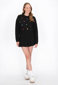 Czarny sweter typu cropped z wielokolorowymi wzorami serc, w połączeniu z czarną spódnicą plisowaną. Obejmuje białe sneakersy i białe skarpety do kolan.