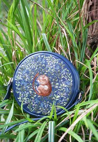 Ümmargune tumesinine kott, millel on tikitud kurrus pruun ahv, ümbritsetud roheliste ja helesiniste lehtedega viinapuudega, puhates kõrges rohus.