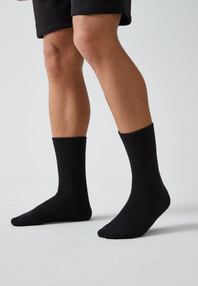 Calcetines de algodón negros de corte crew con textura acanalada, puño ajustado y diseño de puntera sin costuras, usados en pies desnudos, sobre un fondo neutro.
