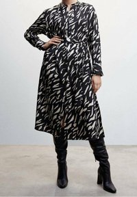 Robe à motif noir et blanc avec des manches longues, une taille cintrée par un nœud et un col boutonné. Le tissu a une texture lisse, avec une fente sur le côté.