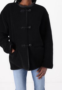 Chaqueta de vellón negra con botones de palanca usada sobre jeans azul claro, manos en los bolsillos, sobre un fondo blanco liso.