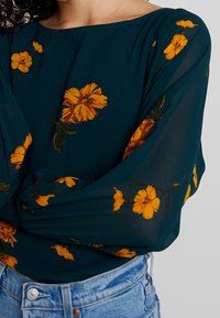Blouse turquoise en tissu léger, ornée de motifs floraux jaunes et à manches longues. Associée à un short en denim bleu taille haute.