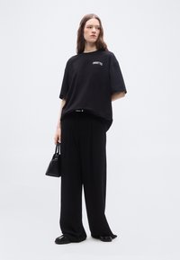 Γυναίκα που φοράει μαύρο φαρδύ oversized μπλουζάκι με το κείμενο "Pirouettes", φαρδιά μαύρα παντελόνια, μαύρα παπούτσια και κρατά μικρή μαύρη τσάντα χειρός, στέκεται σε λευκό φόντο.