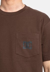 Hnědý bavlněný tričko s kulatým výstřihem, krátkými rukávy a náprsní kapsou s tmavě modrým logem "DC". Hladká textura.