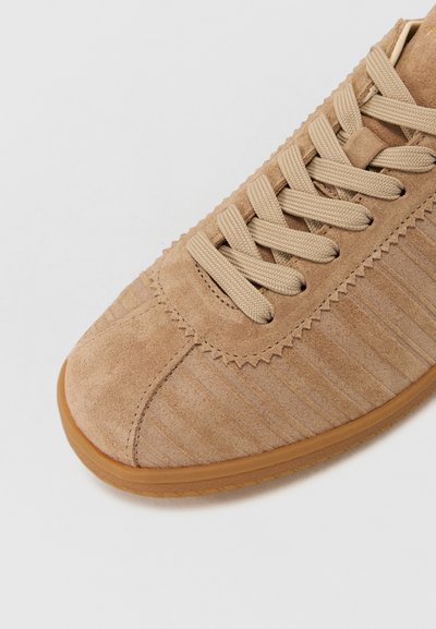 Brūns zamšs sneakers ar ribotām detaļām, noapaļota purngalu, bēšām sieta auklām un brūnu gumijas zoli. Iekļautas zigzaga šuves apmalē.