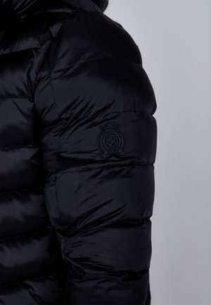 Doudoune noire avec un design matelassé, présentant une surface texturée et un patch logo circulaire sur le haut du bras.