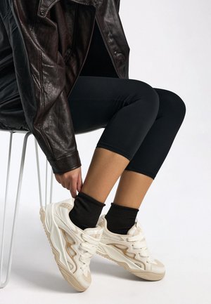Sneakers bianche e beige con dettagli strutturati, indossate con calzini scuri, abbinate a leggings neri e una giacca di pelle marrone su una sedia.