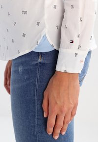 Hvid skjorte med sort bogstav "TH" mønster, lange ærmer, og kontrasterende blå undertrøje synlig. Jeans i denim med subtile slidmærker.
