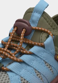 Κοντινό πλάνο ενός παπουτσιού με λουριά σε ανοιχτό γαλάζιο, ελιά υφασμάτινη επιφάνεια, καφέ ελαστικά κορδόνια και καφέ κλειδαριά-κλιπ που ασφαλίζει τα κορδόνια.