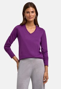 Fioletowa bluzka z długim rękawem z dekoltem w serek, wykonana z miękkiego materiału o gładkiej fakturze i luźnym kroju, zestawiona z jasnoszarymi spodniami.