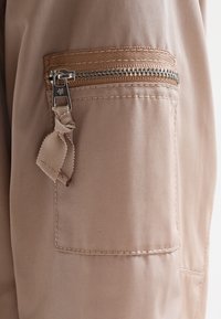 Tela beige con una vista en primer plano de un bolsillo con cremallera, que presenta una cremallera texturizada y una suave cinta como tirador en un diseño de corte recto.