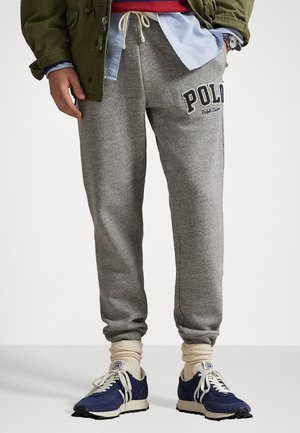 Person wearing gray sweatpants with "Polo" logo, cream socks, and navy blue sneakers, hand in pocket, olive jacket, layered shirts visible.