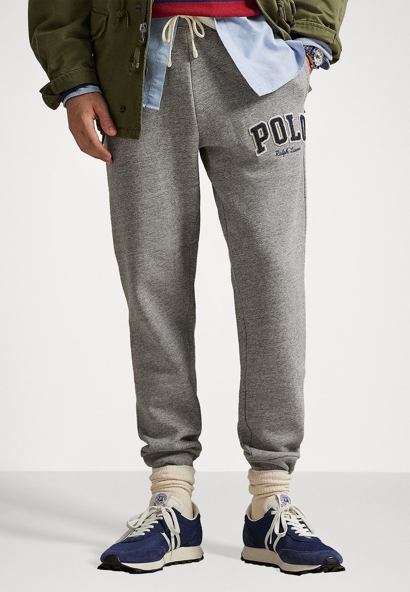 Person, der bærer grå joggingbukser med "Polo" logo, cremede sokker og marineblå sneakers, hånd i lommen, olivengrøn jakke, lagde skjorter synlige.