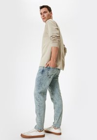 Top de manga larga beige con mangas enrolladas, combinado con jeans de mezclilla azul claro jaspeados y zapatillas blancas con suelas de goma marrón.