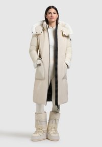 Lange beige pufferjas met een kraag van fauxbont, twee zijzakken, zwarte rits en verstelbare manchetten; gecombineerd met bijpassende laarzen.