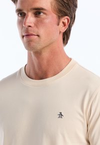 T-shirt beige à manches courtes en tissu doux, avec un col rond et un petit logo de pingouin bleu brodé sur la poitrine.