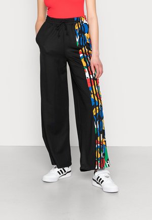 Osoba nosząca czarne szerokie spodnie z kolorowymi abstrakcyjnymi paskami po bokach, białe buty sportowe Adidas z czarnymi paskami oraz czerwony top bez rękawów.