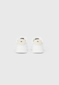 Par bijelih tenisica niskog profila s bež potpeticama i malim crvenim, bijelim i plavim logom straga na neutralnoj sivoj pozadini.