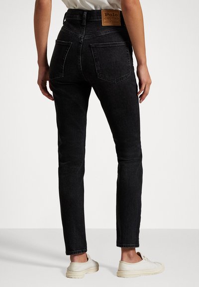 Persona que lleva jeans slim de tiro alto negros con bolsillos traseros y una etiqueta de Polo Denim Goods, combinados con zapatillas blancas.