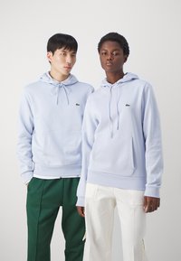 Sweatshirts azul claros com cordões, bolsos frontais e pequenos logotipos. Acompanhados de calças verdes e brancas, com pregas e bolsos.