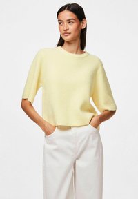 Żółty sweter dzianinowy w prążki z krótkimi rękawami i okrągłym dekoltem, w połączeniu z jasnymi spodniami o luźnym kroju i fakturze.
